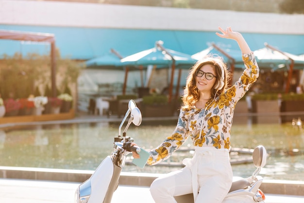 Encantadora chica de pelo oscuro con gafas y auriculares sentado en scooter junto a la piscina al aire libre. Mujer joven con estilo divirtiéndose al aire libre en un día de verano con café en el fondo.