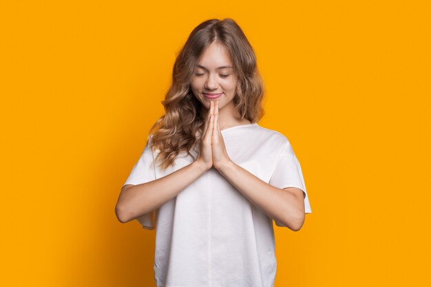 Encantadora chica en ropa casual está orando en una pared de estudio amarilla gesticulando con las palmas y sonriendo con los ojos cerrados