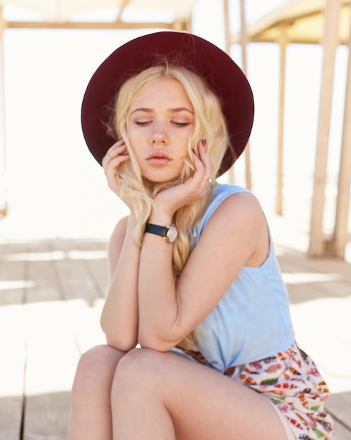 Encantadora chica rubia con cabello rizado, ojos azules, labios grandes y sombrero burdeos posando en el fondo del edificio de madera