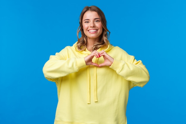 Encantadora chica rubia linda y cariñosa con capucha amarilla que muestra el gesto del corazón y sonríe como simpatía expresa, confiesa en amor o sentimientos conmovedores hacia la persona, pararse en una pared azul