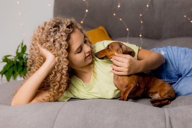 Encantadora chica rubia con rizos está acostada en una cama gris con un perro salchicha