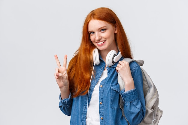 Encantadora colegiala coqueta y descarada pelirroja, usa auriculares sobre el cuello y la mochila, muestra el signo de la paz y sonríe atractiva cámara, de pie feliz sobre blanco disfruta de la vida estudiantil