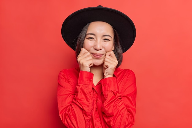 La encantadora dama asiática morena feliz mantiene las manos en las mejillas, sonríe sinceramente, tiene una piel suave y saludable, viste una camisa roja de moda con sombrero negro y posa en el interior. Emociones humanas agradables y expresiones faciales.