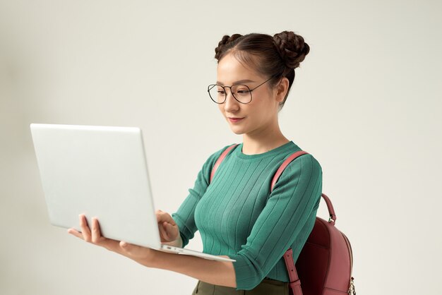 Encantadora dama inteligente está de pie sobre el fondo gris puro con una computadora portátil en sus manos, sonriendo, mirándolo mientras navega, escribe, escribe, usa especificaciones negras