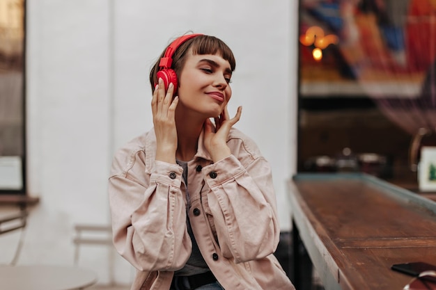 Encantadora dama morena escuchando música afuera Chica elegante con cabello corto en auriculares rojos y traje de mezclilla beige sonriendo al aire libre