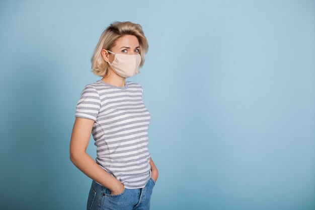 Encantadora dama rubia con una máscara protectora blanca posando en una pared de estudio azul