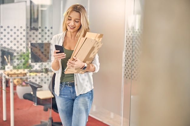 Encantadora dama rubia revisando mensajes en el teléfono inteligente y sonriendo mientras sostiene documentos