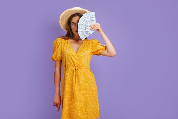 Encantadora dama con sombrero de paja y vestido amarillo de verano mantenga la mitad de la cara cubierta de abanico de efectivo aislado color púrpura ...