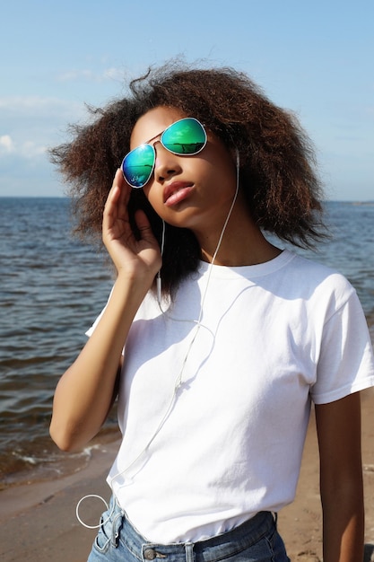 Encantadora e incrível jovem afro-americana em óculos de sol ouvindo música em fones de ouvido em seu celular
