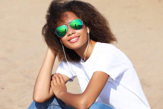 Encantadora e incrível jovem afro-americana em óculos de sol ouvindo música em fones de ouvido em seu celular