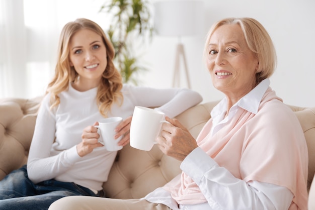 Encantadora y elegante mujer mayor sentada en el sofá junto a su hija, ambas sosteniendo una taza de té y mirando al frente con una sonrisa