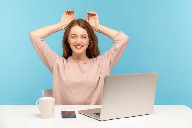 Encantadora empleada joven sonriendo y mostrando orejas de conejo en la cabeza, descansando, divirtiéndose en el lugar de trabajo en la oficina del hogar, sintiéndose despreocupada y feliz. Foto de estudio interior aislado sobre fondo azul.