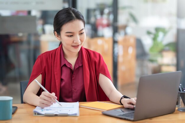 Encantadora empresaria asiática joven en vestido rojo sosteniendo una pluma apuntando a un gráfico usando una computadora portátil en la oficina.