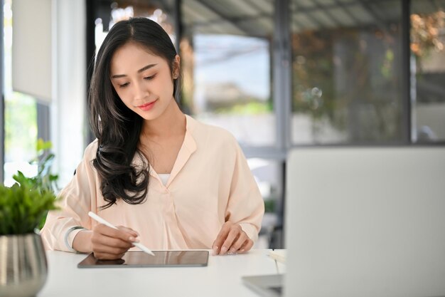 Encantadora empresária asiática milenar com foco em suas tarefas de negócios usando tablet