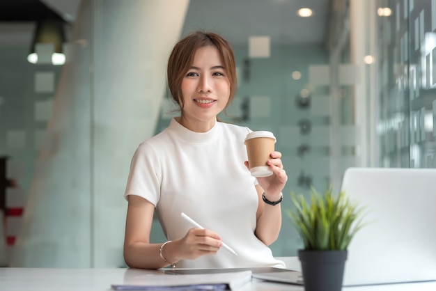 Encantadora empresaria asiática trabaja en tableta listo para beber café concepto de trabajo de oficina. Mirando a la cámara.