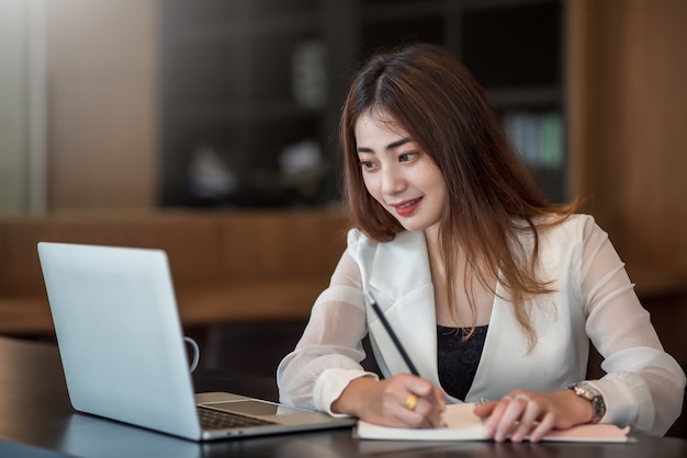 Encantadora empresária asiática trabalhando em um laptop e tomando nota enquanto está sentada à mesa no escritório
