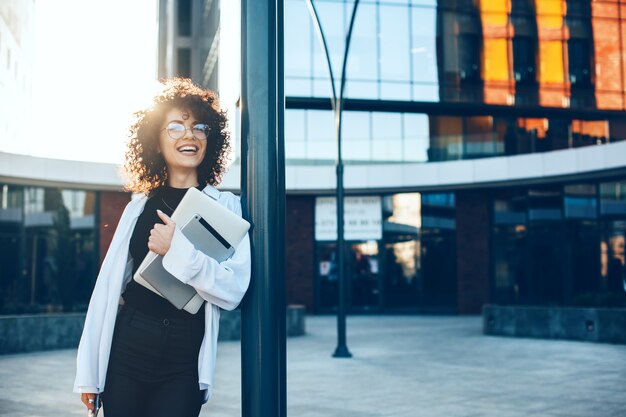 Encantadora empresaria de pelo rizado con gafas sonriendo afuera mientras posa con una computadora portátil y una tableta