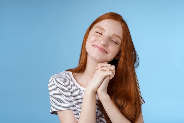 Encantadora encantada feliz soñadora pelirroja novia jengibre cabello largo inclinando la cabeza ojos cerrados sonriendo complacido soñando despierto pensando en dulces recuerdos tiernos presione las palmas juntas, fondo azul.