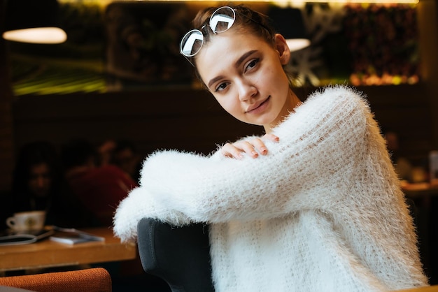 Encantadora estudiante en una elegante blusa blanca se sienta en un restaurante, mira a la cámara y sonríe