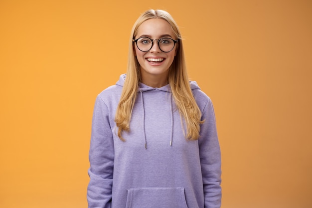 Encantadora estudiante friki sonriente con energía quiere participar en el evento universitario sonriendo mirada emocionada con gafas morado con capucha de pie divertido fondo naranja divirtiéndose. Copia espacio