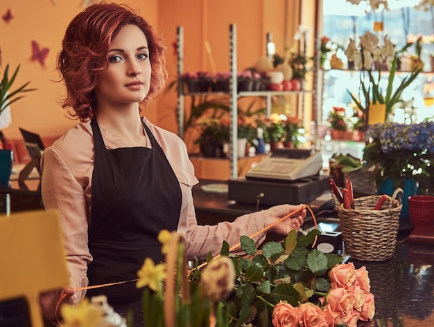 Encantadora floristería pelirroja con uniforme haciendo una hermosa composición floral mientras está de pie en el mostrador de una floristería.