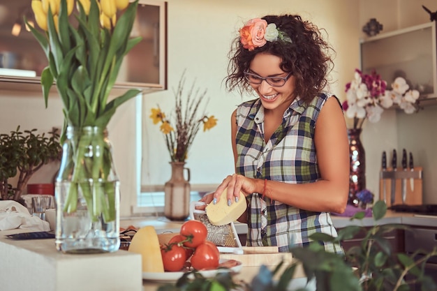 Encantadora garota hispânica encaracolada cozinhando em sua cozinha. Rala o queijo em um ralador.