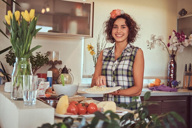 Encantadora garota hispânica encaracolada faz pizza enquanto cozinha em sua cozinha.