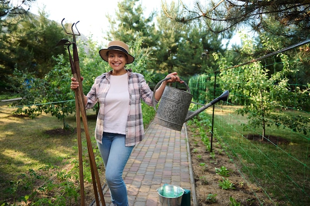 Encantadora jardinera posa para la cámara con herramientas de jardinería regadera y azada contra un fondo de granja orgánica