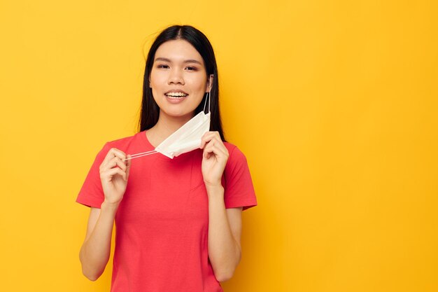 Encantadora jovem asiática em camiseta vermelha proteção de máscara médica fundo amarelo inalterado