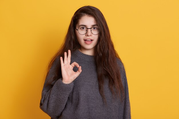 Encantadora jovem de cabelos escuros veste camisola preta mostrando sinal de ok com os dedos