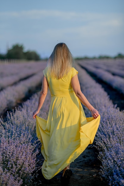 Encantadora jovem em campos florescentes de lavanda Vista traseira da adorável senhora usando chapéu de vestido amarelo acenando óleo essencial de lavanda com flores frescas de lavanda conceito de massagem spa aromaterapia