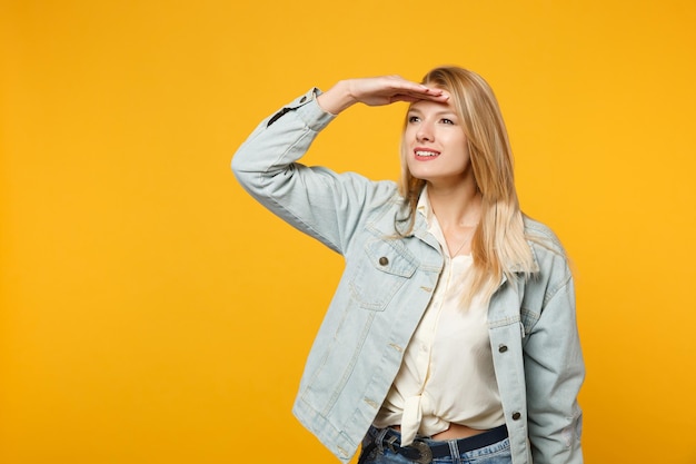 Encantadora jovem em roupas casuais jeans, segurando a mão na testa, olhando de lado a distância distante isolada no fundo da parede laranja amarela no estúdio. Conceito de estilo de vida de pessoas. Mock up espaço de cópia.