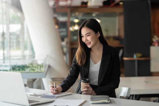 Encantadora jovem empresária asiática tomando notas com o laptop no escritório.