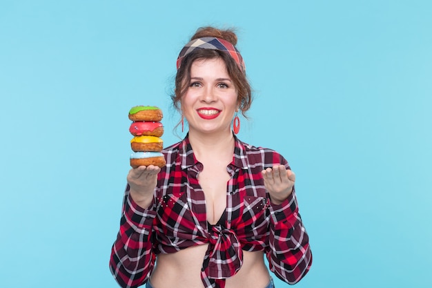 Encantadora jovem garota pin-up segurando dois donuts nas mãos e um posando sobre um fundo azul. Benefícios do conceito e vendas de doces e panificação. Copyspace