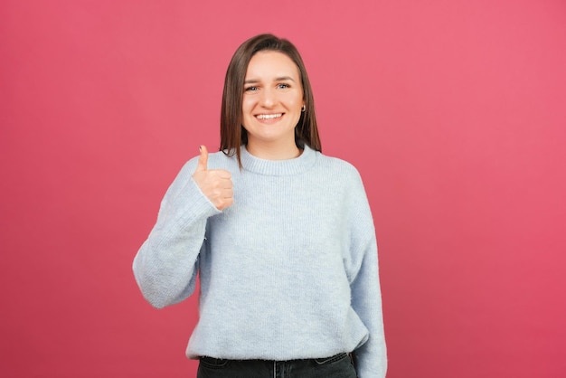 Encantadora jovem sorridente está segurando o polegar sobre fundo rosa