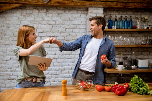 Encantadora joven alegre cocinando la cena juntos, mirando la receta en la tableta digital y divirtiéndose en la cocina rústica