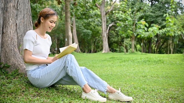 Encantadora joven asiática o estudiante universitaria sentada debajo de un árbol y leyendo un libro