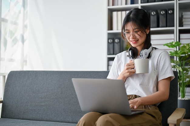 Una encantadora joven asiática usa su computadora portátil mientras toma café en un sofá en su sala de estar