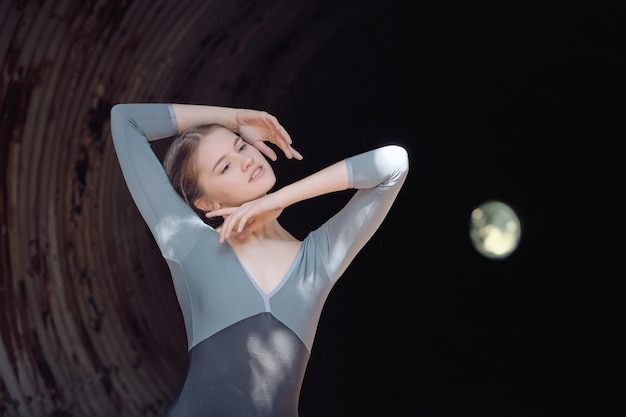 Encantadora joven bailarina de ballet delgada con los brazos en la cabeza mirando a lo lejos mientras ensaya en un largo túnel oscuro con paredes destartaladas