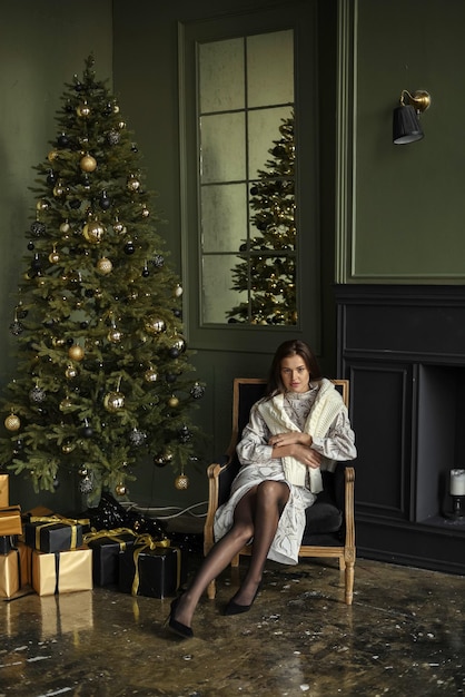 Foto una encantadora joven se está calentando junto a la chimenea con un suéter cerca del árbol de navidad