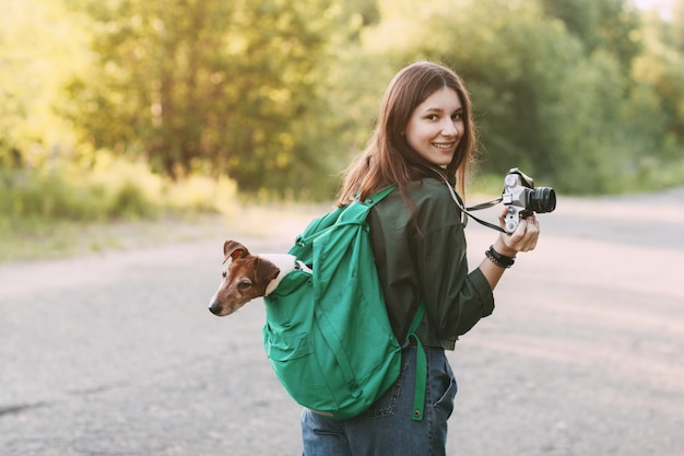 Una encantadora joven camina en la naturaleza, sosteniendo una mochila en su hombro, desde la cual mira su perro, y sosteniendo una cámara en sus manos.