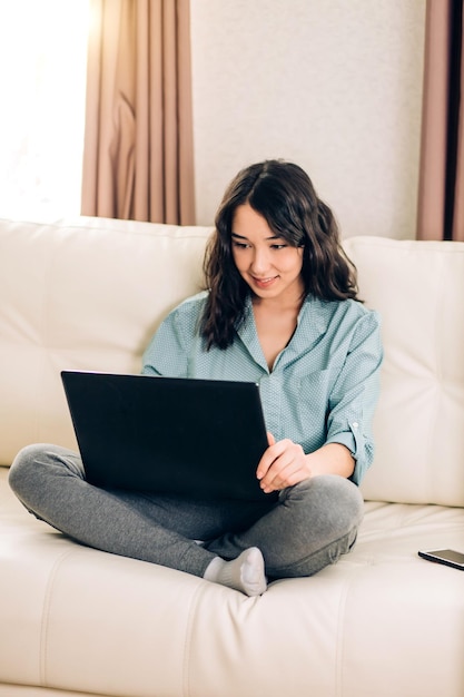 Encantadora joven caucásica vestida con ropa doméstica informal, sonriendo mientras ve videos, chateando en línea con amigos en una computadora portátil negra sentada en un acogedor sofá en un apartamento moderno.