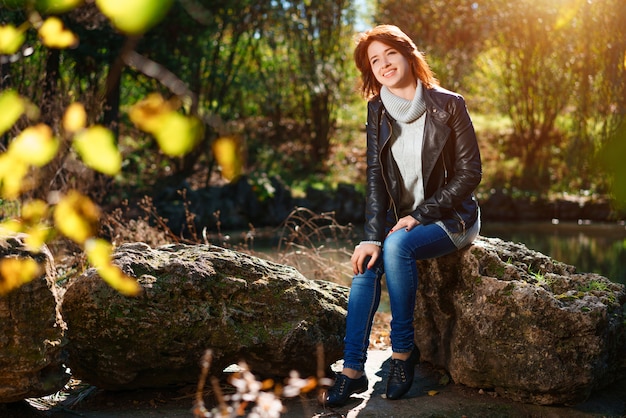 Una encantadora joven está sentada en una roca junto a un estanque en un parque de otoño en el sol