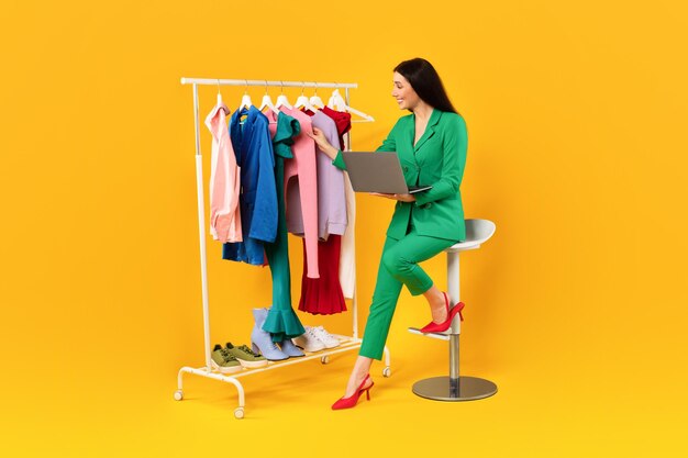 Una encantadora joven europea con una computadora portátil tocando la ropa en el estante eligiendo y comprando en línea nuevas