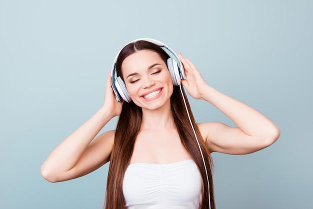 Encantadora joven con gran sonrisa en auriculares modernos. Ella es soñadora y feliz, escucha su canción favorita, sostiene los auriculares, con los ojos cerrados en el espacio puro.