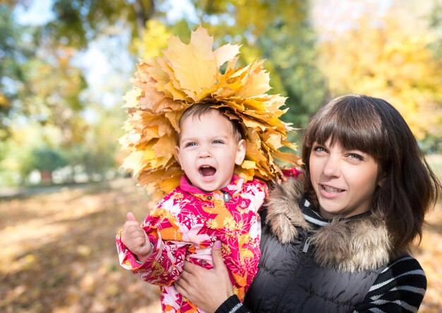 Encantadora joven madre sostiene en sus brazos a una niña bonita con una corona de hojas de otoño de arce amarillo en la cabeza mientras camina en el parque. Concepto de otoño