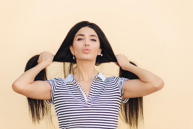 Encantadora joven mujer caucásica muestra su largo cabello oscuro y recto, los labios muestran poses de belleza natural contra un fondo claro vestida con una camiseta a rayas Concepto de belleza y cuidado del cabello