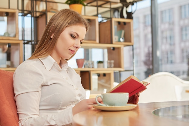 Encantadora joven mujer de negocios leyendo un libro, descansando en la cafetería