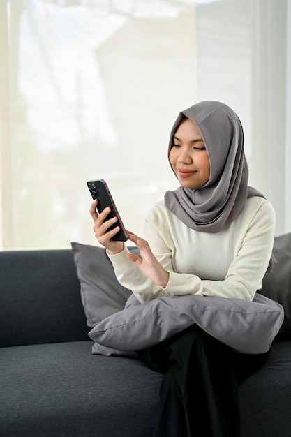 Encantadora joven musulmana asiática usando hiyab usando su teléfono inteligente mientras se relaja en el sofá