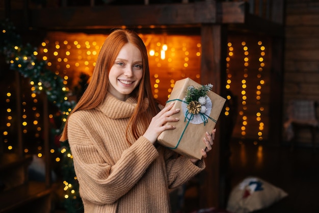 Encantadora joven pelirroja sosteniendo una hermosa caja de regalo de Navidad en el fondo de luces borrosas